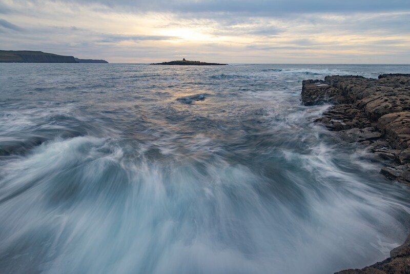 Waves surge across a rocky coastline at dusk with headlands visible across calm waters.