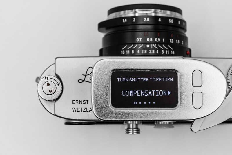 Top-down view of a rangefinder camera with lens mounted, displaying shutter compensation menu on LCD screen.