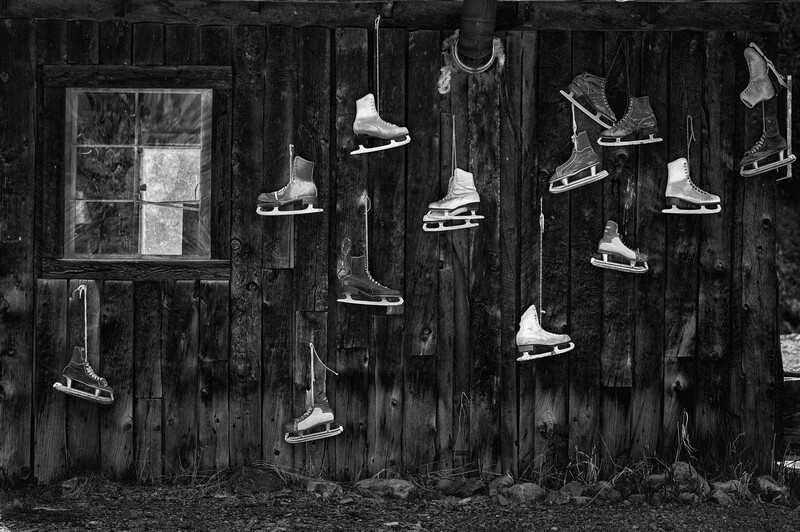Multiple pairs of ice skates suspended by their laces against a weathered wooden wall with a small window.