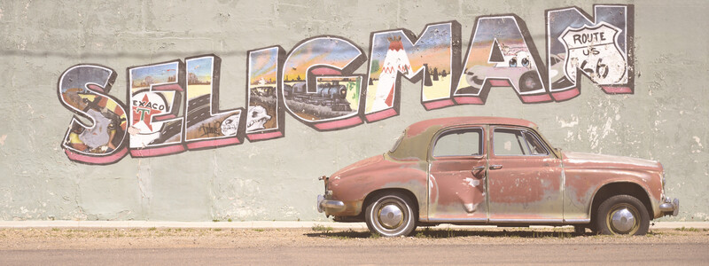 Rusty 1950s sedan parked in front of Route 66 mural on weathered concrete wall.