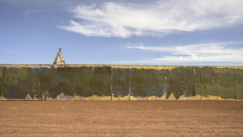 Earthen fortification wall with wooden palisade extending across arid landscape, featuring a pyramidal structure visible above the rampart.