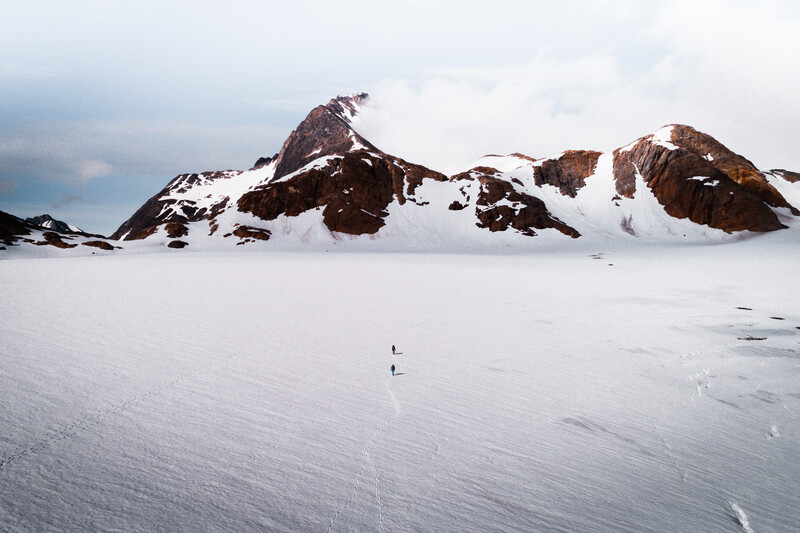 Lone figure standing on a vast snow-covered landscape with rocky, snow-capped peaks rising behind.