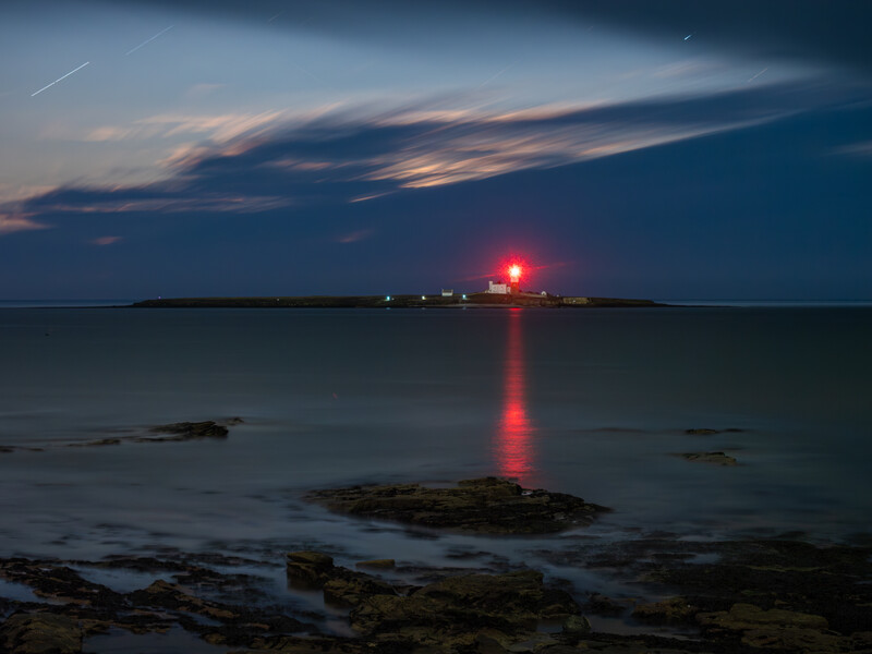 Red lighthouse beacon illuminated at dusk with rocky shoreline in foreground and dark water reflecting the light.