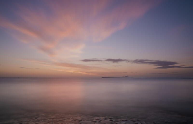 Calm seascape at dusk with soft pink and purple clouds reflected in still water.
