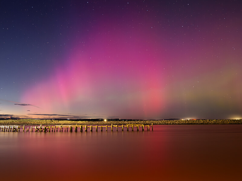 Aurora borealis display over a coastal pier with wooden pilings and distant buildings reflected in calm water.