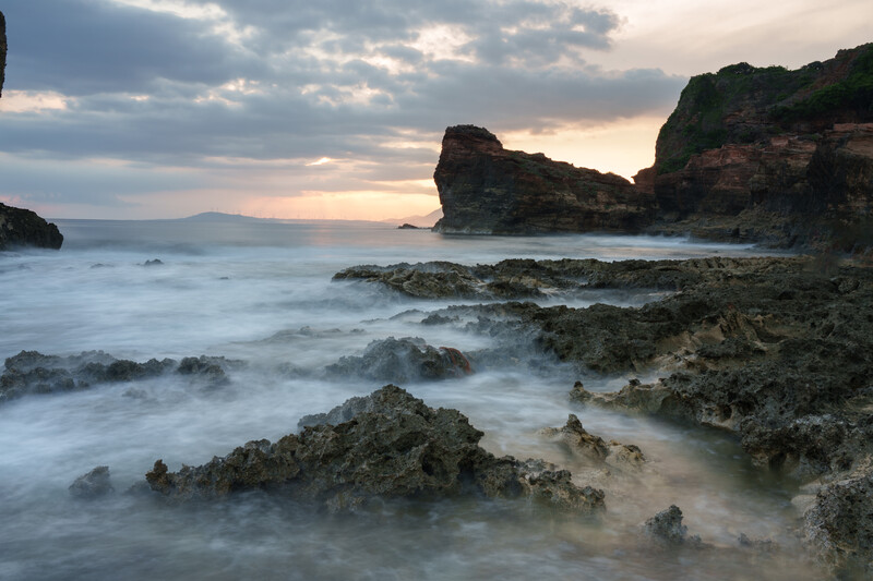 Rocky coastline at dusk with smooth water and dramatic cliff formations backlit by sunset sky.
