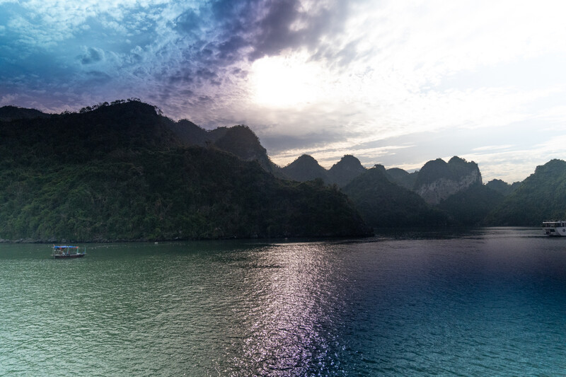 Limestone karst mountains rise dramatically from turquoise water at sunset with moored boats and golden light reflecting across the bay.