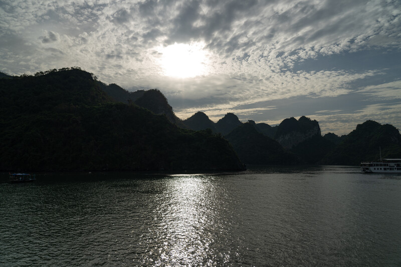 Silhouetted limestone karst formations across calm water under dramatic cloudy sky with sun breaking through.