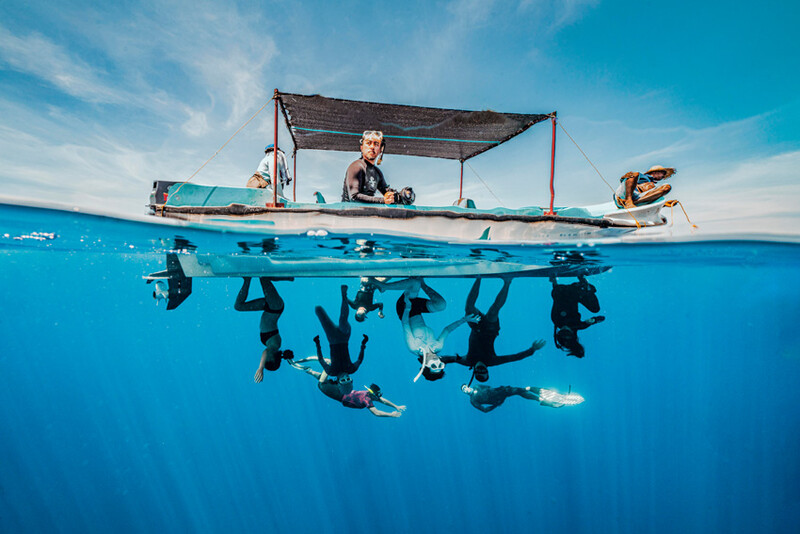 Split-level photograph showing a boat operator above water and multiple divers submerged below, framed by the water's surface.