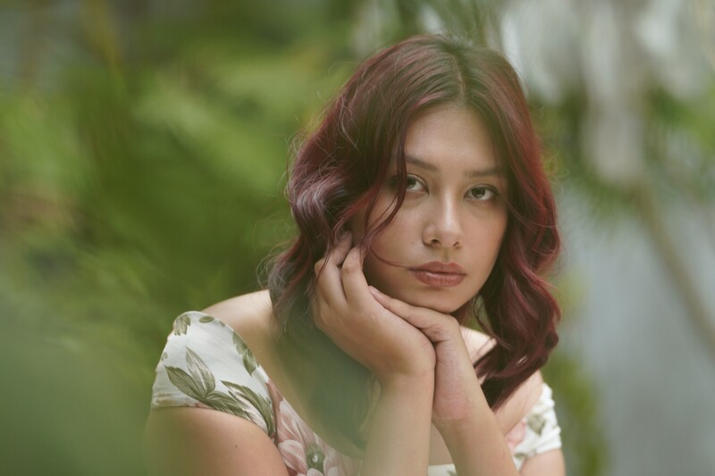 Young woman with reddish-brown wavy hair resting her chin on her hand, gazing thoughtfully to the side.