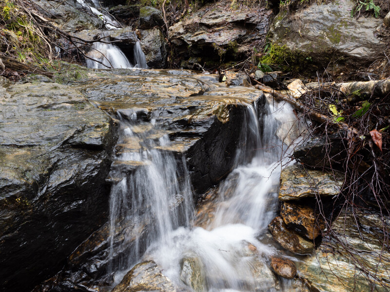 Cascading waterfall flowing over layered slate rocks with moss and vegetation on surrounding banks.