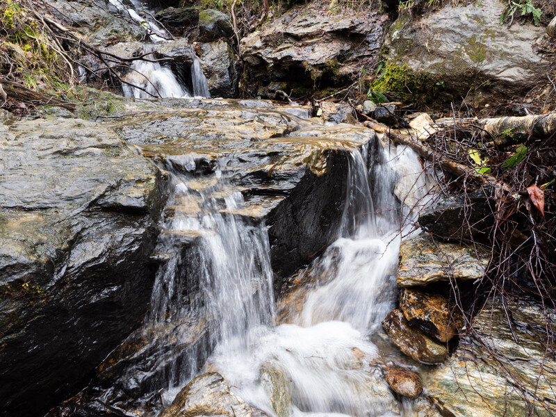 Multi-tiered waterfall cascading over layered slate rock formations surrounded by moss and vegetation.