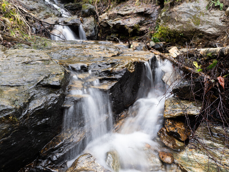 Multi-tiered waterfall cascading over layered slate rocks surrounded by moss and vegetation.