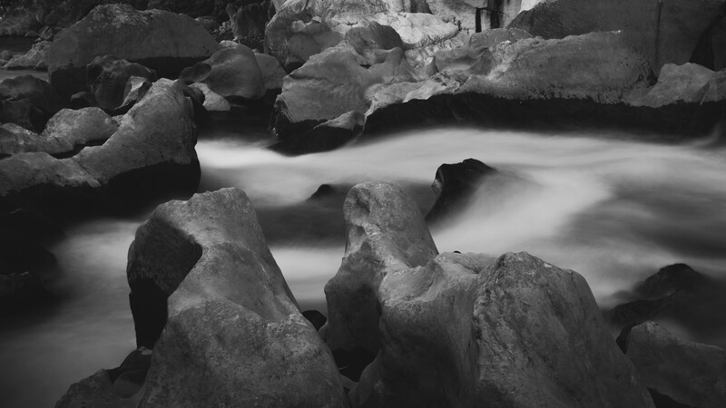 Flowing stream water cascading smoothly around large boulders in black and white.