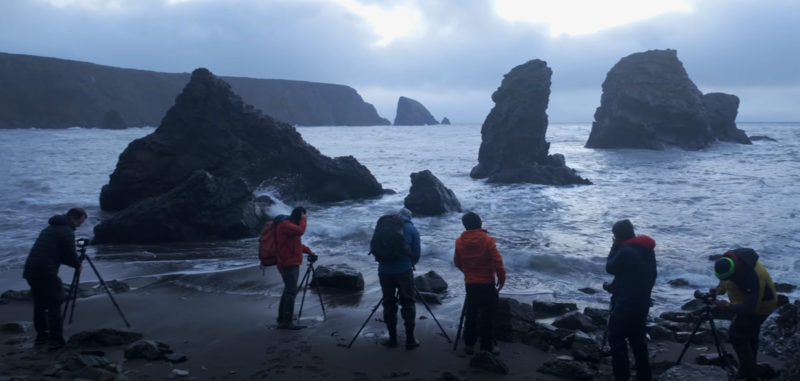 Group of photographers with tripods on a dark sand beach, shooting dramatic rocky sea stacks under overcast sky.