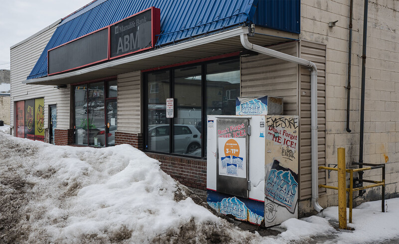 Weathered convenience store with snow-covered entrance and graffitied ice cooler in winter light.