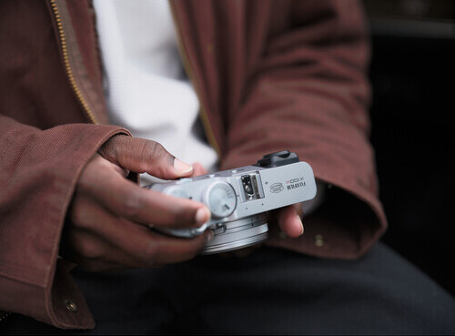 Photographer holding a compact silver camera with their hands while wearing a burgundy jacket.