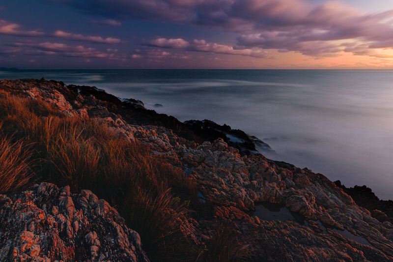 Rocky coastal hillside with golden grasses overlooking calm ocean at dusk.