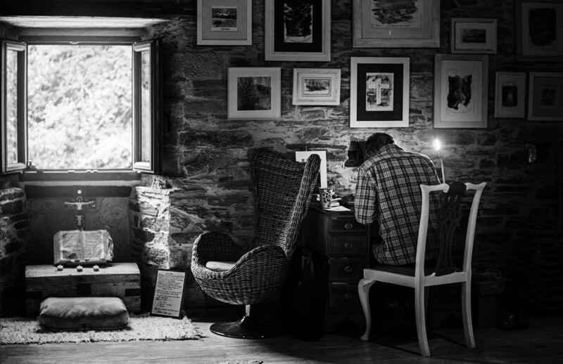 Two people seated at a desk in a stone-walled room, viewed from behind in black and white photography.