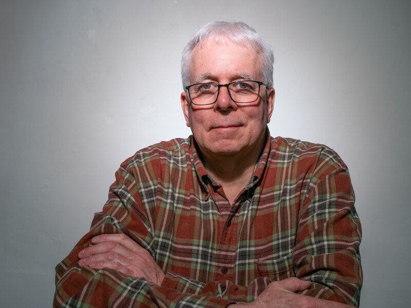 Older man with white hair and glasses wearing a red and green plaid shirt in a studio portrait.
