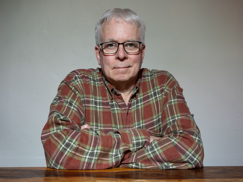 Older man with gray hair and glasses wearing a red plaid shirt, seated at a wooden table against a neutral background.