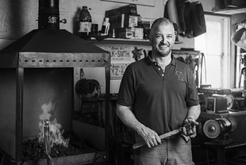 Photographer smiling in studio holding light meter, with kiln or furnace visible on left and equipment on shelves behind.