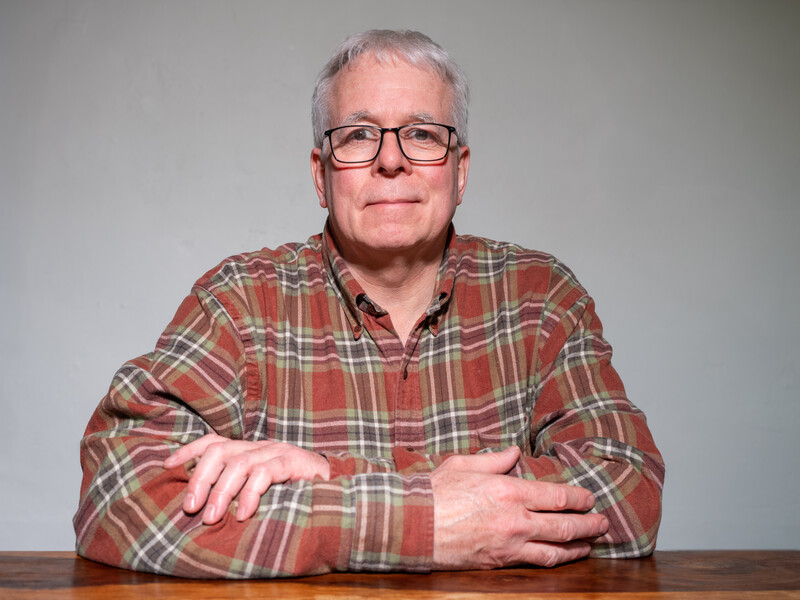 Older man with gray hair and glasses wearing a red plaid shirt, seated at a wooden table against a neutral background.