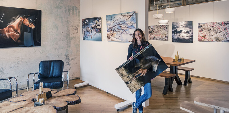 Woman holding a large framed photograph in a gallery space with artwork on white walls.