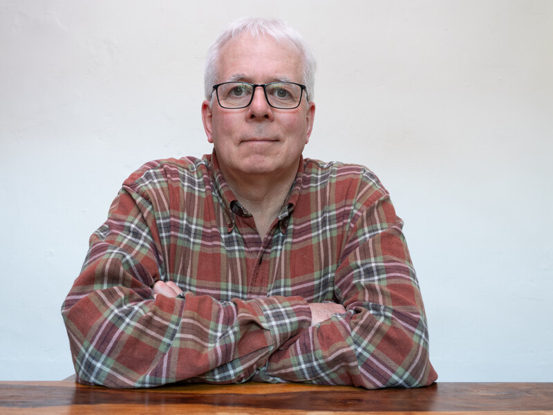 Man with white hair and glasses wearing a red plaid shirt, seated at a wooden table against a white background.