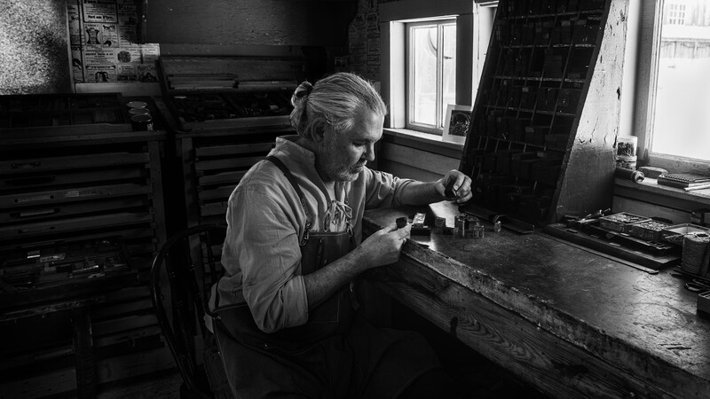 Older craftsman working at a bench in a dimly lit workshop, focused on detailed handiwork.