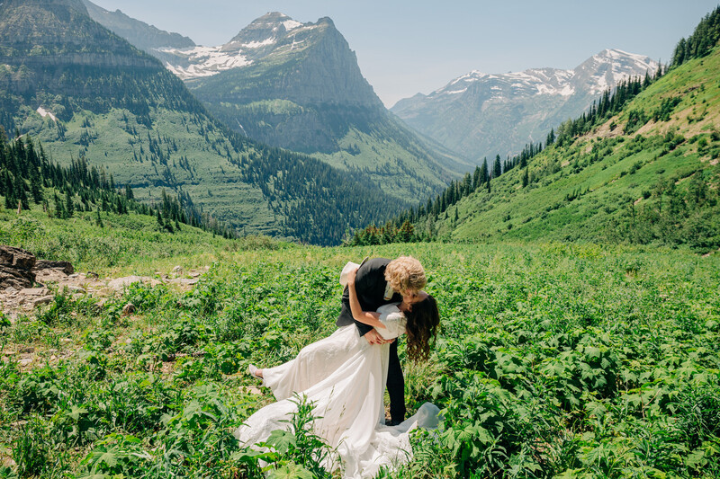 Bride and groom lean for a kiss in a lush green valley under hard light