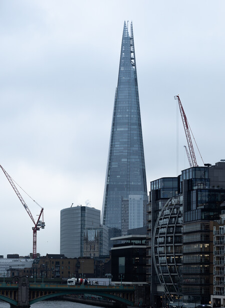 Tall tapered glass tower rises above surrounding London riverside buildings under overcast sky.