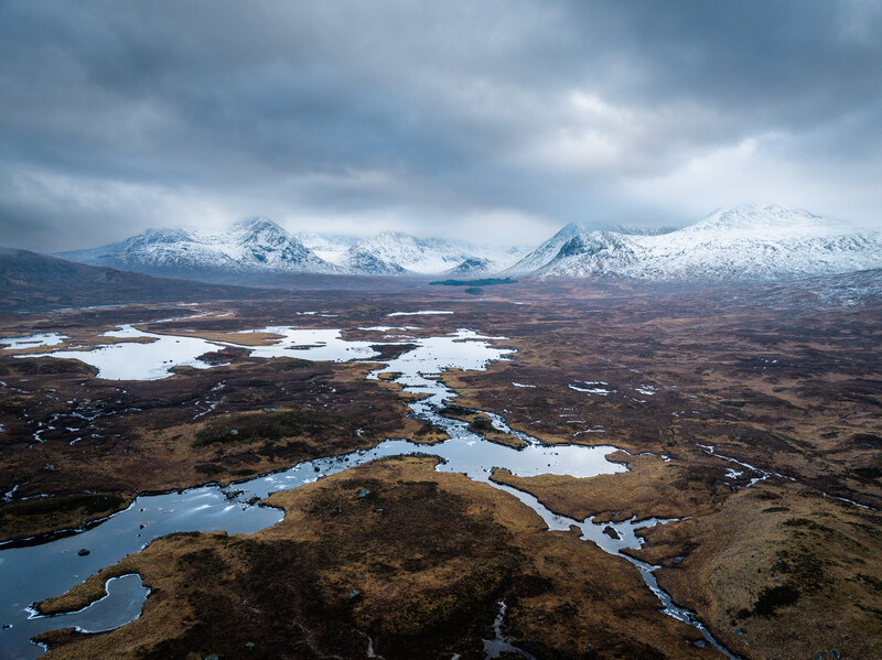 Aerial landscape of snow-capped mountains, scattered blue lakes, and brown tundra terrain under dramatic clouds.