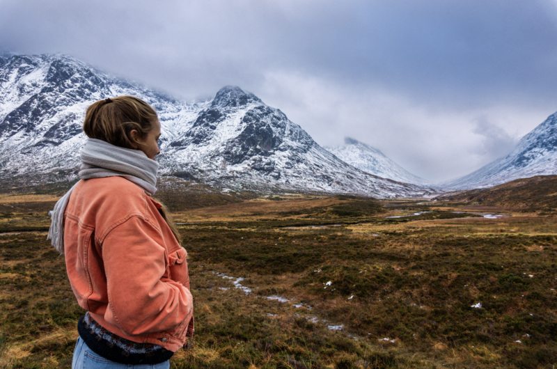 Person in coral jacket gazing toward snow-capped mountains across tundra landscape.