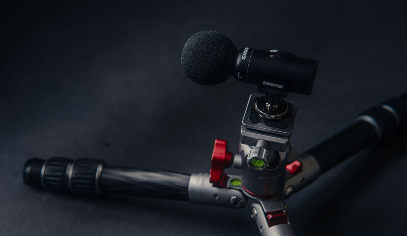 Professional video rig with fluid head, red handles, and shotgun microphone mounted on top against dark background.