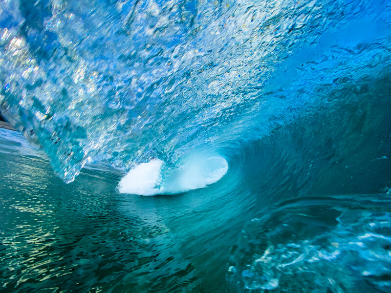 Underwater view looking up through a wave barrel toward bright sky and surface light.