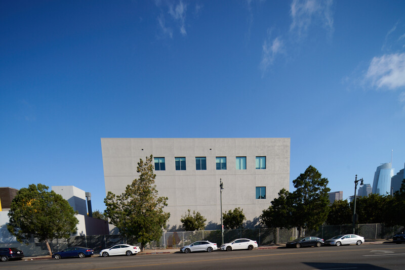 Modern cubic building with cream-colored facade and grid of blue-framed windows, parked cars in foreground.