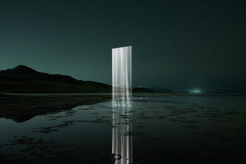 Illuminated vertical rectangular structure reflected in still water at night with mountains and starry sky in background.