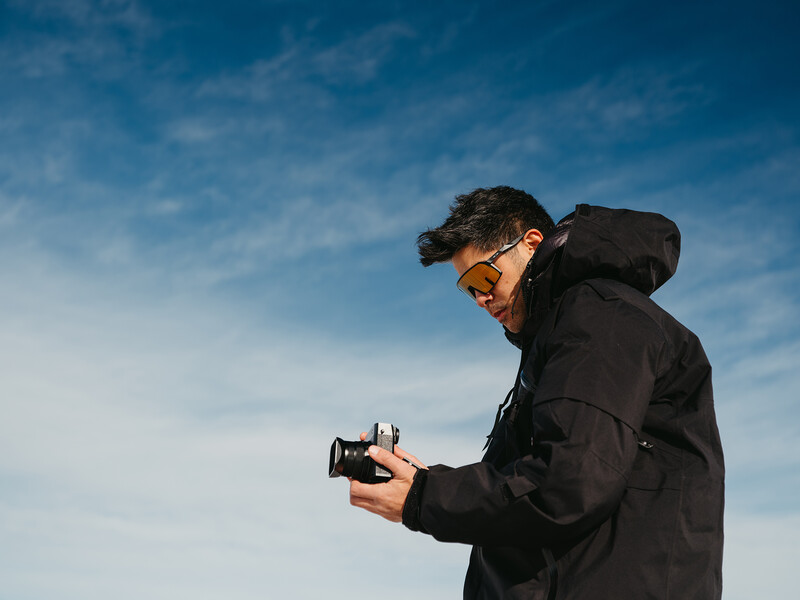 Photographer wearing sunglasses and black jacket holding a vintage camera against a clear blue sky.