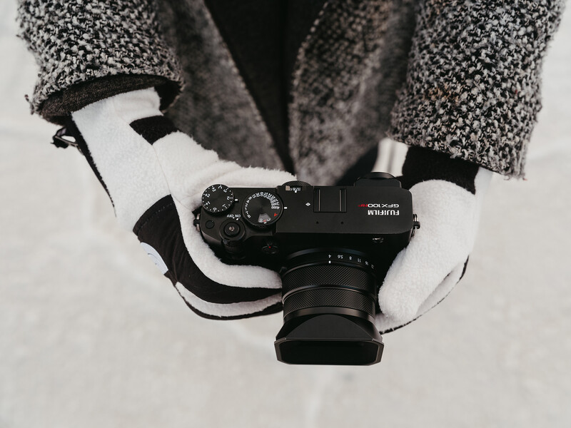 Photographer wearing white gloves holding a black rangefinder camera against a textured gray jacket.