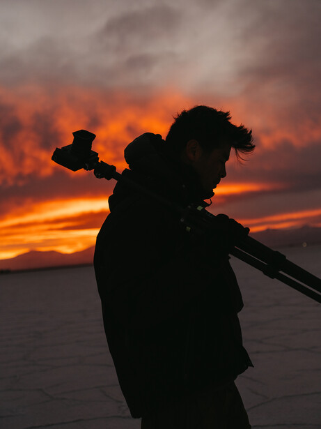 Silhouetted photographer holding a camera with telephoto lens against a dramatic orange and pink sunset sky.
