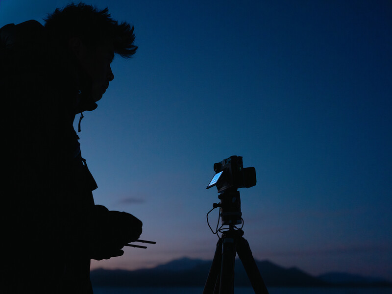 Silhouetted photographer operating a camera on a tripod during twilight, with a person watching from the foreground.