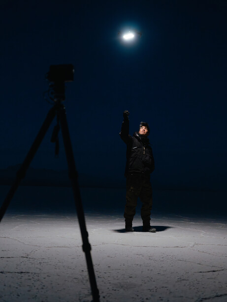 Man in dark clothing standing on pavement at night, illuminated by a bright light source mounted on a tripod above.