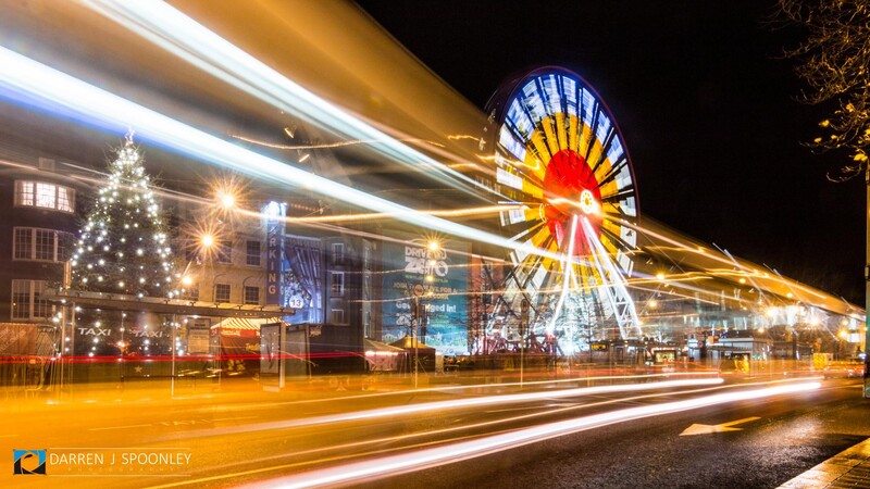 Illuminated Ferris wheel and Christmas tree at night with light trails from passing traffic.