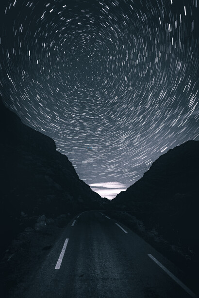 Long exposure photograph of stars spiraling above a dark road stretching toward a mountain pass.