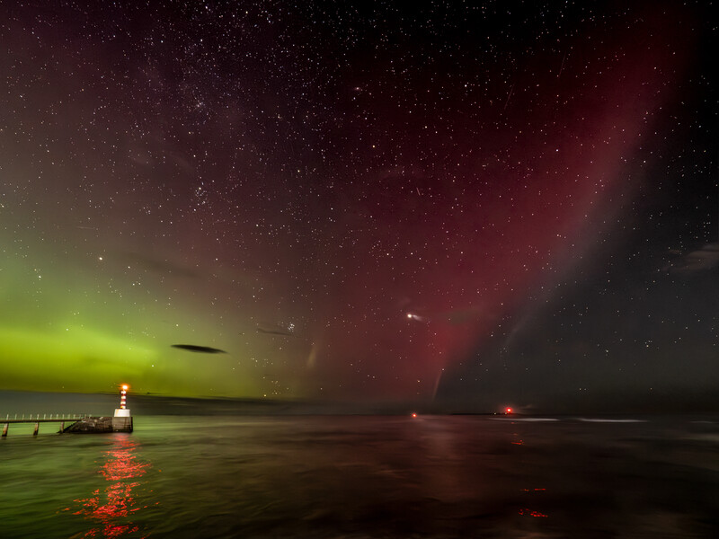 Aurora borealis illuminating a dark sky in green and red hues above a calm ocean with a distant lighthouse.