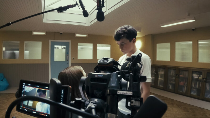 Cinematographer operating a professional video camera while seated actress performs on set in studio with glass-walled offices and skylighting.