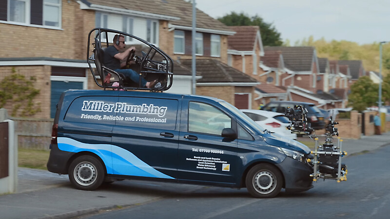 Blue and navy plumbing service van with operator visible on roof-mounted lift platform in residential street.
