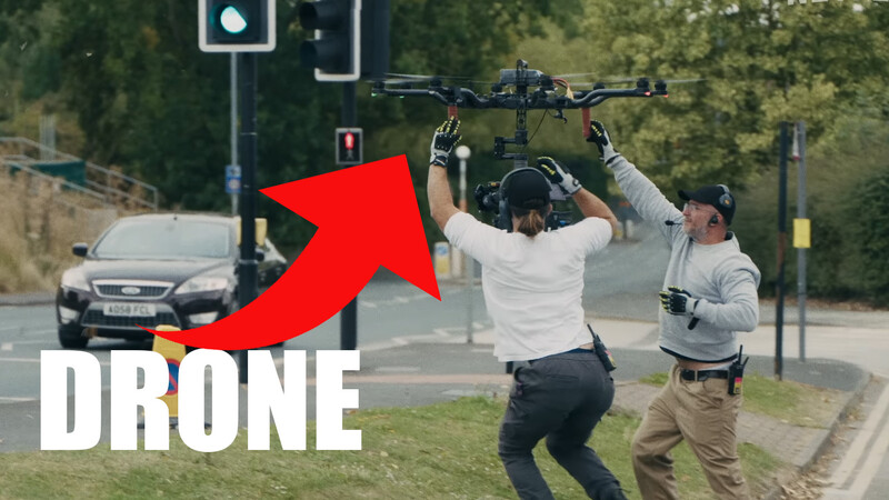 Two people lifting a drone overhead on a city street with traffic light and parked car visible in background.