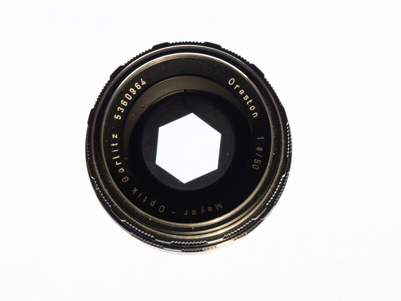Vintage camera lens with hexagonal aperture opening and brass-colored markings on black body.